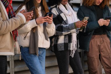 Öğrenciler duvarın yanında duran akıllı telefonları kullanıyor, kışlık kıyafetler giyiyorlar.