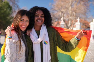Eşcinsel onur geçidinde sarılıp gökkuşağı bayrağı tutan iki etnik kadın, lgbtq artı cemaati kutluyor.