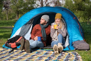Mutlu çift ormanda kamp yaparken çadırın içinde rahatlıyor ve sıcak içecek içiyor.