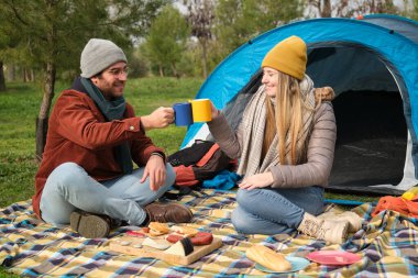 Genç çift, dağlarda kamp yaparken çadırlarının önünde kahvaltı ederken kadeh kaldırıyor.