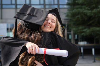 İki bayan mezun birbirine sarılıyor, akademik başarılarını neşe ve diplomayla kutluyorlar.
