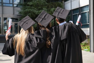 Üç mezun akademik önlük giyiyor ve ellerinde diplomayla üniversite binasının yakınında birbirlerine sarılıyorlar.