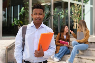 Üniversite binasının önünde elinde kitap ve sırt çantası olan gülümseyen bir üniversite öğrencisinin portresi. Arka planda sohbet eden iki kız.