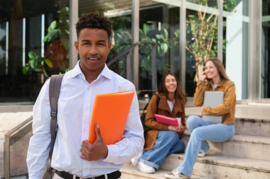 Elinde turuncu bir klasör olan gülümseyen bir üniversite öğrencisinin portresi. Arka planda sınıf arkadaşları oturuyor.