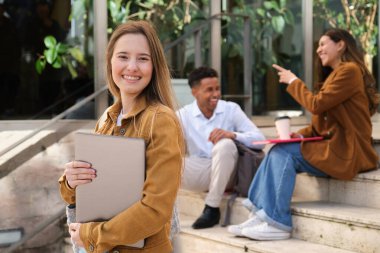 Sınıf arkadaşları konuşurken ve merdivenlerde oturmuş eğlenirken elinde dizüstü bilgisayar tutan gülümseyen bir kız öğrencinin portresi.