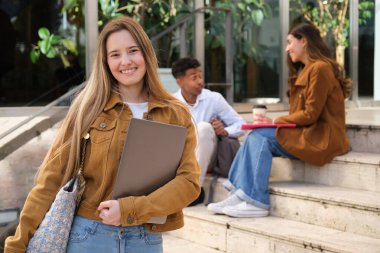 Elinde dizüstü bilgisayarı olan gülümseyen bir kız öğrencinin portresi üniversite merdivenlerinde duruyor ve arka planda iki öğrenci oturuyor ve konuşuyor.