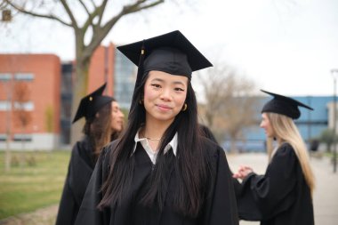 Üniversite kampüsünde mezuniyet elbisesi ve şapkasıyla gülümseyen Asyalı genç mezun.