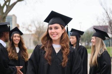 Mezuniyet elbisesi ve şapkası giymiş neşeli bir üniversite öğrencisinin portresi. Kampüste arkadaşlarıyla kutlama yapıyor.