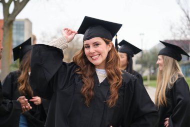 Mezuniyet şapkasını düzelten neşeli bir üniversite öğrencisinin portresi, kampüste sınıf arkadaşlarıyla kutlama yapıyor.