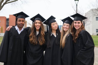 Mezuniyet kıyafetleri ve şapkalar giyen beş neşeli etnik öğrenci kampüste birlikte poz veriyorlar.