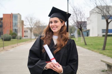 Üniversite kampüsünde gülümseyen ve diplomasını tutan genç bir mezun.