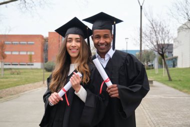 Mezuniyet elbisesi giyen ve üniversite kampüsünde diploma taşıyan gülümseyen iki etnik öğrenci.