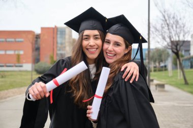 Mezuniyet gününde birbirlerine sarılmış, gülümseyen ve diplomalarını tutan iki kadın mezunlar.