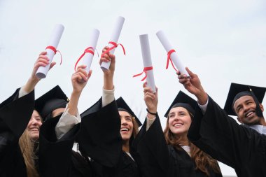 Bir grup neşeli mezun, önlük ve şapka giyip diplomalarını havaya kaldırarak akademik başarılarını kutluyorlar.