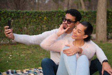 Genç Latin çift parkta akıllı telefonlarıyla selfie çekiyor, komik suratlar yapıyorlar.
