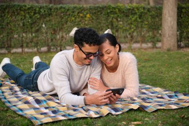 Genç, çok ırklı bir çift parkta rahatlatıcı bir günün tadını çıkarıyor. Battaniyeye uzanıp akıllı telefonlarıyla sosyal medyayı izliyorlar.