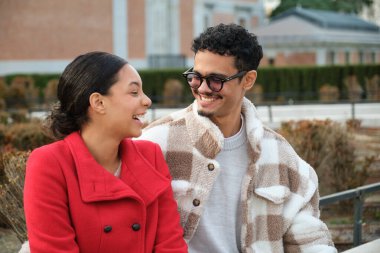 Parktaki bankta oturmuş gülen ve konuşan çok ırklı genç bir çift.