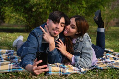 Genç çift parkta bir battaniyeye uzanıp akıllı telefonlarıyla selfie çekiyorlar, dillerini çıkarıp komik suratlar yapıyorlar.