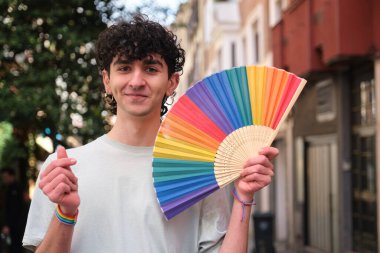 Gökkuşağı yelpazesi tutan genç adam geçit töreninde kolunda lgbt bilekliğiyle kalp işareti yapıyor.