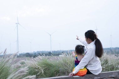 Beautiful young mother daughter relax sitting summer meadow background and windmill,Wind turbines are alternative electricity sources, the concept of sustainable resources, People in the community with wind generators turbines, Renewable energy.