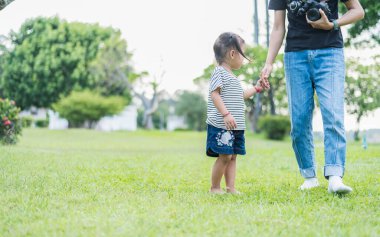 Annenin küçük bir çocukla el ele tutuşup yeşil çimlerde yürüdüğü yakın plan. El ele tutuşan anne ve kız, siyah saçlı Asyalı kız..