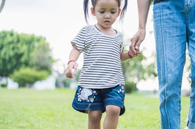 Annenin küçük bir çocukla el ele tutuşup yeşil çimlerde yürüdüğü yakın plan. El ele tutuşan anne ve kız, siyah saçlı Asyalı kız..