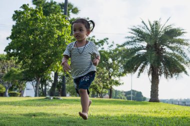 Parkta yeşil çimlerde koşan mutlu kız. Parktaki çayırda güneş ışığı altında neşeli çocuklar. Çocuk bahar çimlerinde oynuyor, genç kadın akşamları koşuyor..