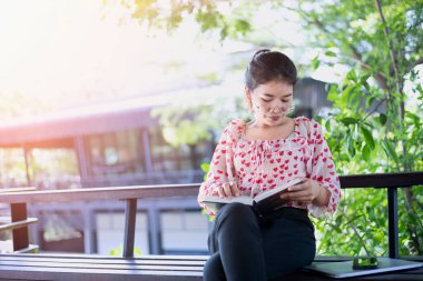Koyu renk saçlı, gözlüklü Asyalı kadın bir bankta oturur ve evinin önündeki çardakta kitap okur..