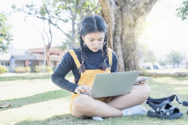 Genç kız öğrenci yeşil çimlerin üzerinde dizüstü bilgisayarla parkta oturuyor. Açık havada okuyan öğrenciler alanı kopyalayın.