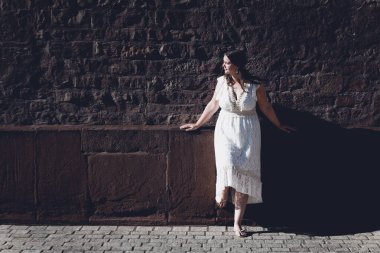 Beyaz elbiseli bir kadın tuğla duvarın önünde duruyor.