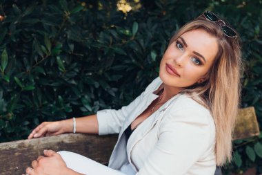 Beyaz ceketli ve güneş gözlüklü bir kadın bankta oturuyor.