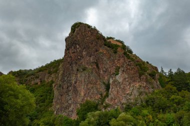 Kayalık yüzeyi ve birkaç ağacı olan bir dağ. Gökyüzü bulutlu ve dağ yeşil bitkilerle kaplı.