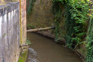 Paslı bir boru, yeşil sarmaşıklarla kaplı iki taş duvarın arasından geçen dar ve sığ bir kanala akar. Kanal beton ve yosunla kaplı..