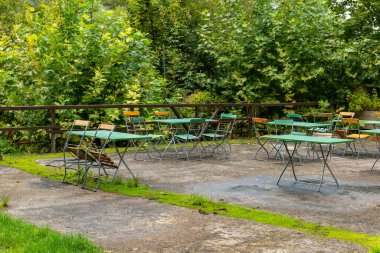 Çakıl taşı yüzeyi olan bir veranda yeşil ağaçlar ve çalılarla çevrilidir. Metal masalar ve yeşil kapaklı sandalyeler verandanın etrafına saçılmış..