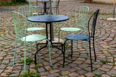 Dört sandalyeli yuvarlak siyah bir masa küçük, kırmızımsı kahverengi kaldırım taşlarından yapılmış bir verandada oturur. Sandalyelerin hassas siyah çerçeveleri ve yeşil koltukları var. Sandalyeler masanın etrafına dizildi..