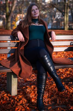 Parktaki bankta uzun bir palto ve uzun botlar giymiş bir kadın, canlı sonbahar yaprakları gibi açık gökyüzünün altındaki zemini örtüyor..