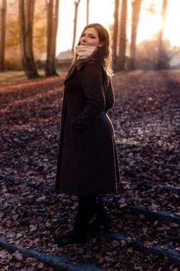 Bir kadın renkli sonbahar yapraklarıyla kaplı bir yolda yürüyor. Koyu renk bir ceket ve eşarp giyer, gün batımında huzurlu manzaranın tadını çıkarır..