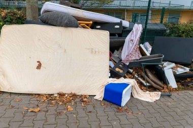Bir döşek, sandalye ve kutular da dahil olmak üzere bir yığın mobilya kaldırıma saçılmış ve etrafı dökülen yapraklarla çevrilmiş, bölgede devam eden atık sorunlarını vurguluyor.