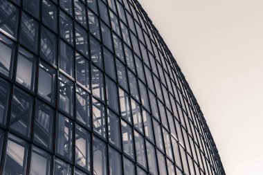 Gösterişli bir mimari tasarım, ışığı yansıtan cam panellerden yapılmış eğimli bir cephe sergiliyor, şehir ortamında modern şehir tasarımının özünü yakalıyor..