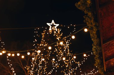 Sıcak ışık dizileri bir kış gecesini aydınlatıyor, çok güzel dekore edilmiş bir Noel ağacını parlayan bir yıldızla süslüyor. Neşeli atmosfer neşeyi ve kutlamayı davet eder..