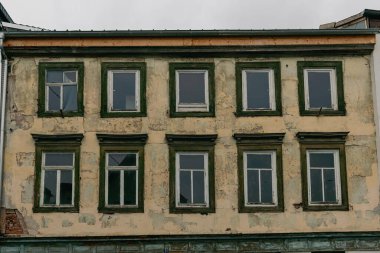 Eski bir binanın yıpranmış cephesinde soyulmuş boyalı birkaç pencere bulunur. Yapı, bulutlu bir gökyüzüne karşı kurulmuş yaş ve ihmal tarihini yansıtıyor..