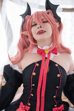 Bir manken, çarpıcı siyah ve kırmızı bir kıyafet giymiş canlı pembe saçlı bir kostüm karakteri sergiliyor. Hava parlak ve davetkar.