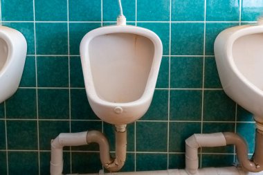 Üç beyaz pisuvar halka açık bir tuvaletin parlak turkuaz döşeli duvarına karşı dizilmiş. Doğal ışık boşluğu aydınlatır, temiz su tesisatını aydınlatır.