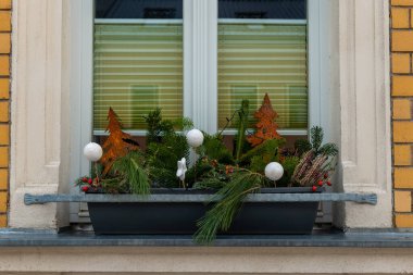 Dekoratif bir vitrin kutusunda kış yeşilliği, çam dalları ve şenlikli süsler sergileniyor. Sahne tatil sezonunda rahat bir şehir atmosferini yansıtıyor.