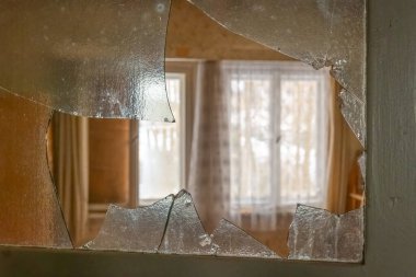 Kırık bir cam levha, perdeleri geri çekilmiş, hafifçe aydınlatılmış bir odayı gösteriyor. Toz ve durgunluk boşluğu doldurur, unutulmuş hikayeleri ima eder.