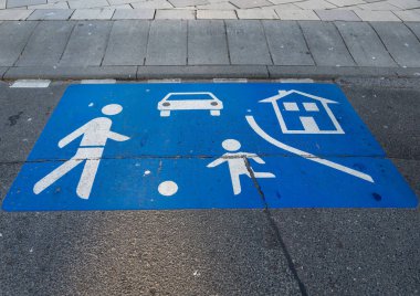 Mavi yol işaretinde bir yayanın, bir çocuğun, bir arabanın ve bir evin sembolleri yer alıyor ve şehir ortamındaki ailelerin güvenliğini vurguluyor. Bu tasarım trafiğin yanında tedbirli davranışı teşvik eder..