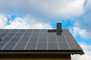 Güneş panelleri, güneş ışığını aktif olarak bulutlu mavi gökyüzü altında enerjiye dönüştüren meskun bir çatının bir kısmını kaplar ve yenilenebilir enerji kullanımını gösterir..