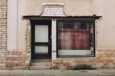 Yıpranmış bir binanın ön cephesinde tuğlalar ve perdeli geniş bir pencere bulunuyor. Giriş, süslenmemiş, sakin bir sokakta yaşı ve sadeliği yansıtıyor..