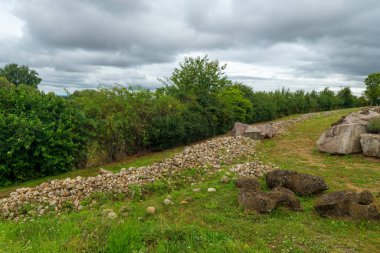 Yıpranmış bir taş duvar, arka planda sık ağaçlar bulunan yemyeşil bir manzaranın yanında uzanır. Gökyüzü gri bulutlarla dolu, bu da potansiyel yağmuru gösteriyor..