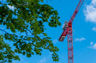 Uzun kırmızı bir vinç parlak mavi bir gökyüzüne karşı duruyor, yemyeşil yapraklarla çevrili. Sahne, gün ışığında kentsel gelişimi gösteriyor.